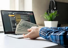 Person holding cash beside a laptop displaying financial data on screen.