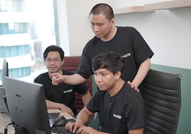 A group of LevelUp team members collaborating in an office, reviewing data on a computer screen, and discussing financial strategies while wearing branded company shirts.