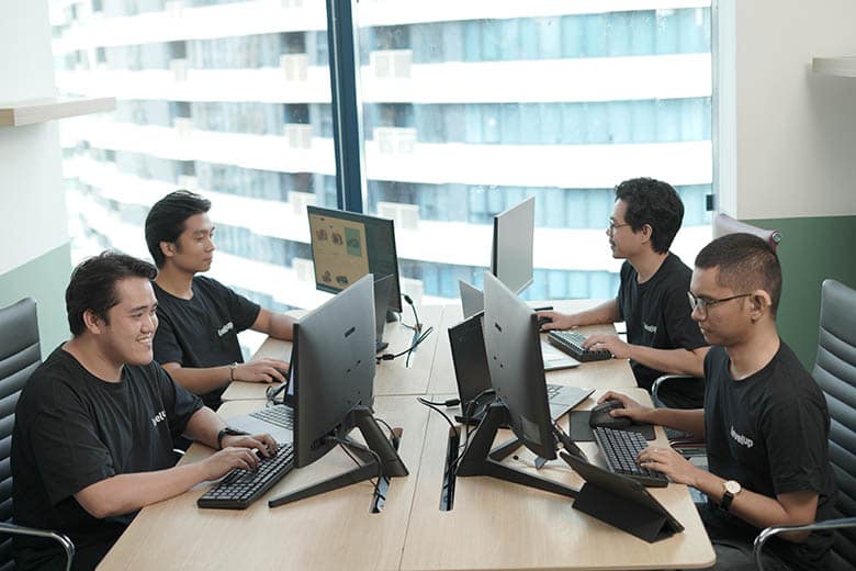 Four individuals seated at a modern office table, working on desktop computers. They are wearing matching black T-shirts and appear to be engaged in tasks on their screens.