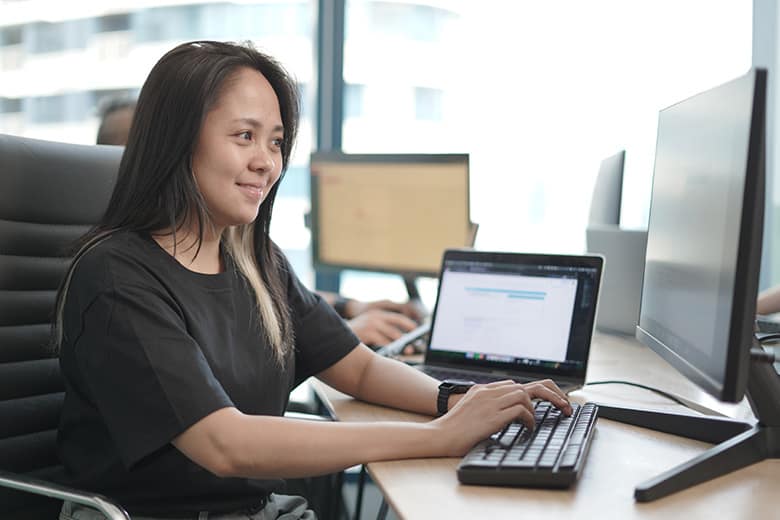 A female personal assistant working on a computer. She is smiling and appears focused. She is wearing a black T-shirt, and there are multiple monitors and a laptop in front of her.