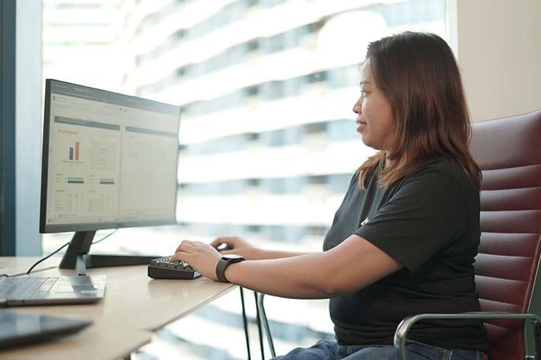 GLB06506 - optimized A professional woman in a black t-shirt sits at a computer workstation, viewing data analytics or performance metrics displayed on a monitor.