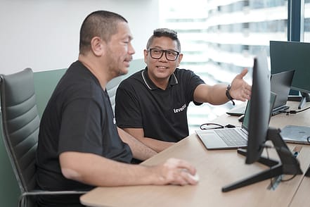 Two men collaborating at a desk, both wearing black shirts. One man gestures towards the computer screen while the other listens attentively. A laptop and other office items are visible on the desk.