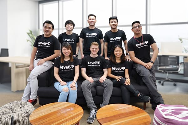 A group photo of LevelUp team members, smiling and posing in front of a bright office space, all wearing LevelUp shirts with a view of office furniture in the background.