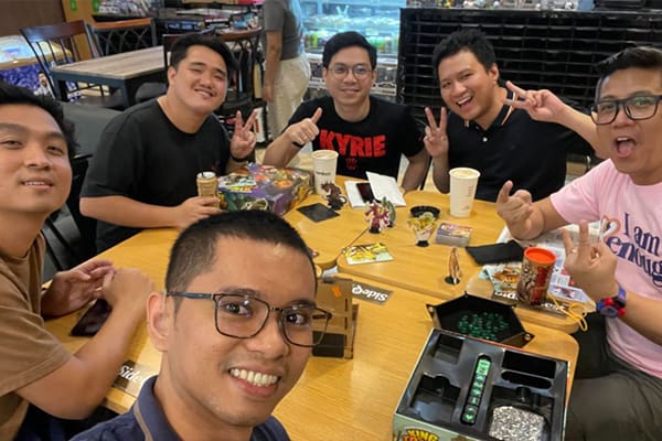 Group of people playing board games and posing for a selfie at a table with various game pieces and drinks in the foreground.