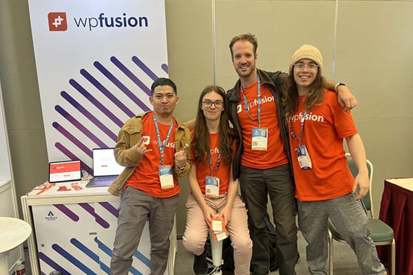 Four people in orange T-shirts, smiling together at the WP Fusion booth during WordCamp Asia.