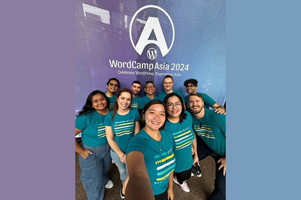 Group of professionals in WordCamp Asia 2024 t-shirts smiling together in front of a colorful banner with WordCamp Asia 2024 branding.