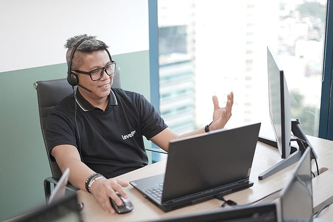 A telesales specialist wearing a headset and LevelUp-branded polo shirt, engaging in a virtual call at his workstation with multiple monitors.