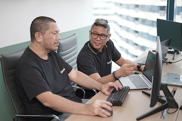 Two telemarketers wearing LevelUp-branded shirts working together at a modern office setup, discussing strategies while using a desktop and laptop.