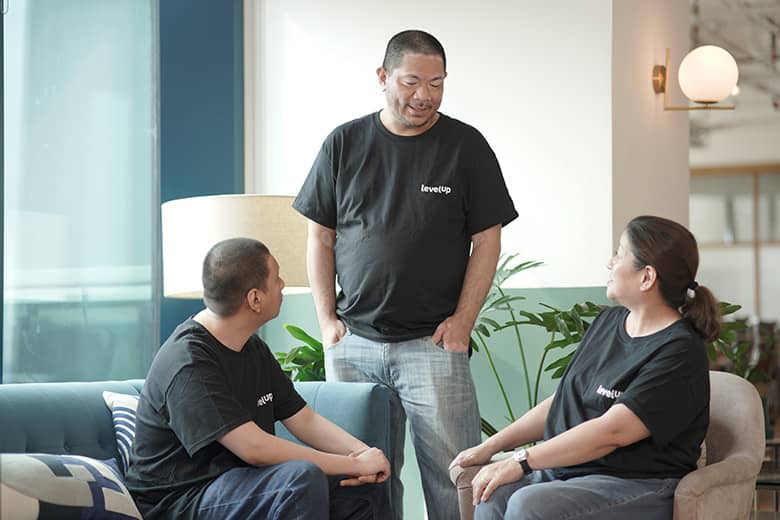 Three professionals having a conversation, with two seated on a couch and one standing nearby, engaging in discussion.