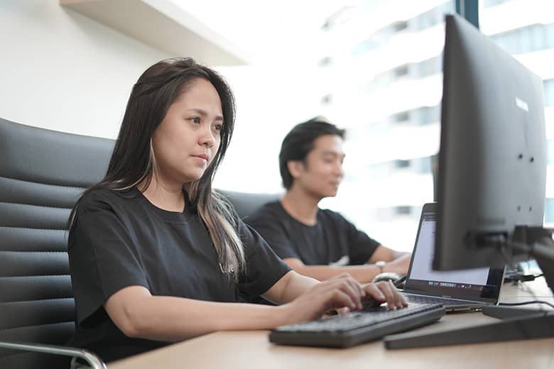 A woman in a t-shirt focused on typing at her desk, with a colleague in the background.