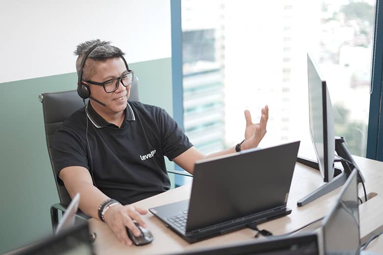 Sales Support Specialist wearing a headset, working at a desk with multiple monitors and a laptop, engaged in a video or phone conversation in a modern office setting.