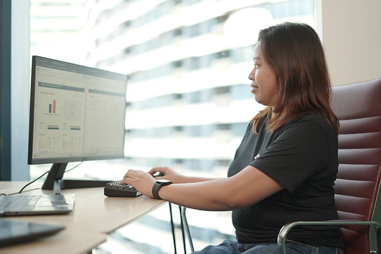 A professional woman in a black t-shirt sits at a computer workstation, viewing data analytics or performance metrics displayed on a monitor.