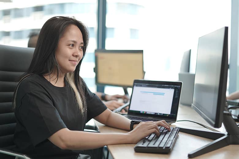 A smiling woman in a black t-shirt types on a keyboard with a laptop and monitor in front of her, in a modern office setting.