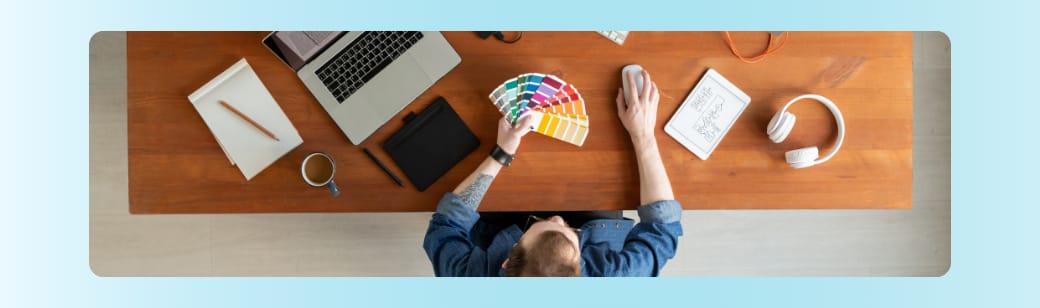 A designer sitting at a desk with color swatches, tablet, headphones, and laptop, preparing creative materials.